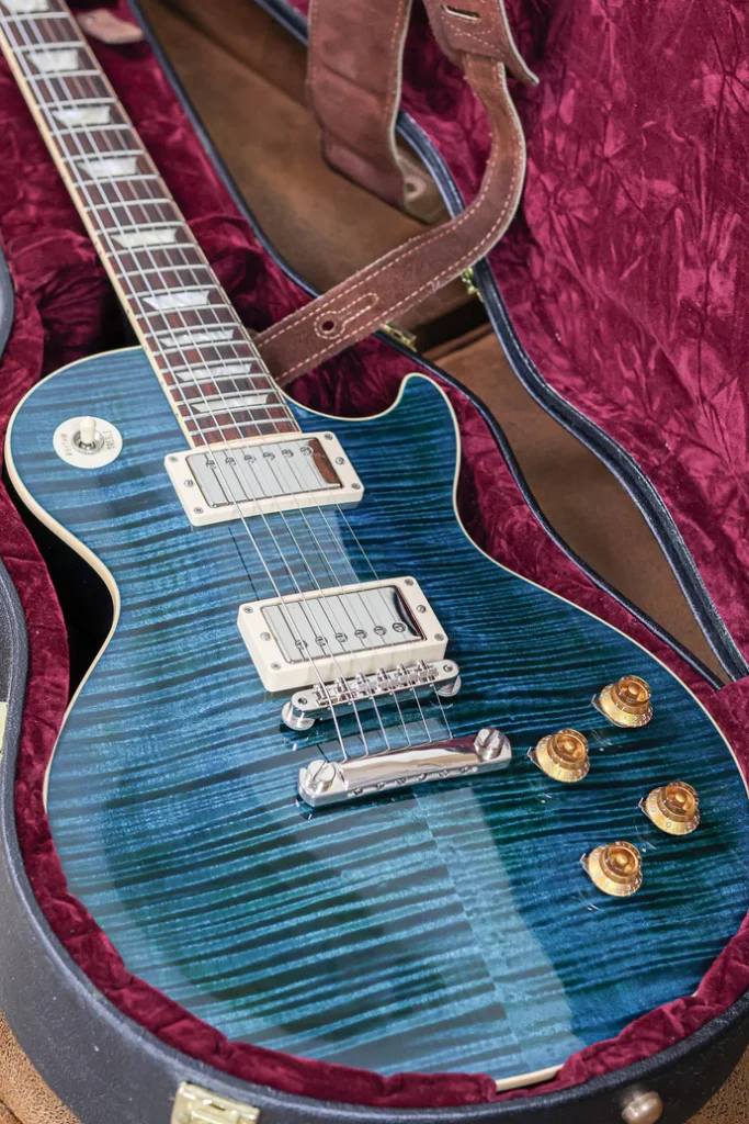 Gibson Les Paul electric guitar in translucent blue finish with flamed maple top resting inside a plush red velvet hardshell case with a brown leather strap.
